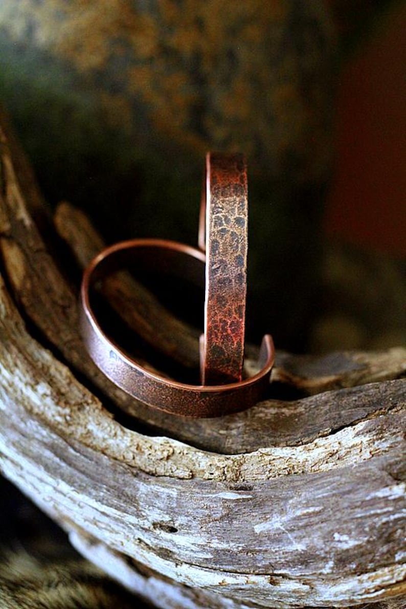 May include: Two copper-colored cuff bracelets with a textured finish. One bracelet is standing upright, while the other is lying down. The bracelets are displayed on a piece of weathered wood, showcasing their rustic appeal.
