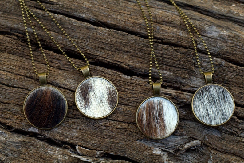May include: Four necklaces with round pendants featuring a cowhide pattern. The pendants are a light brown and white colour and are set in a bronze-coloured metal frame. The necklaces are on a wooden surface.