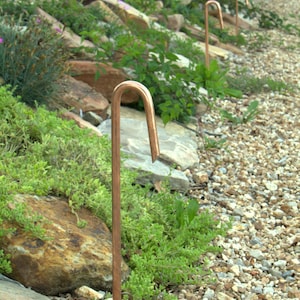 May include: A row of copper garden path lights are installed in a gravel pathway. The lights are curved at the top and are set into the ground.