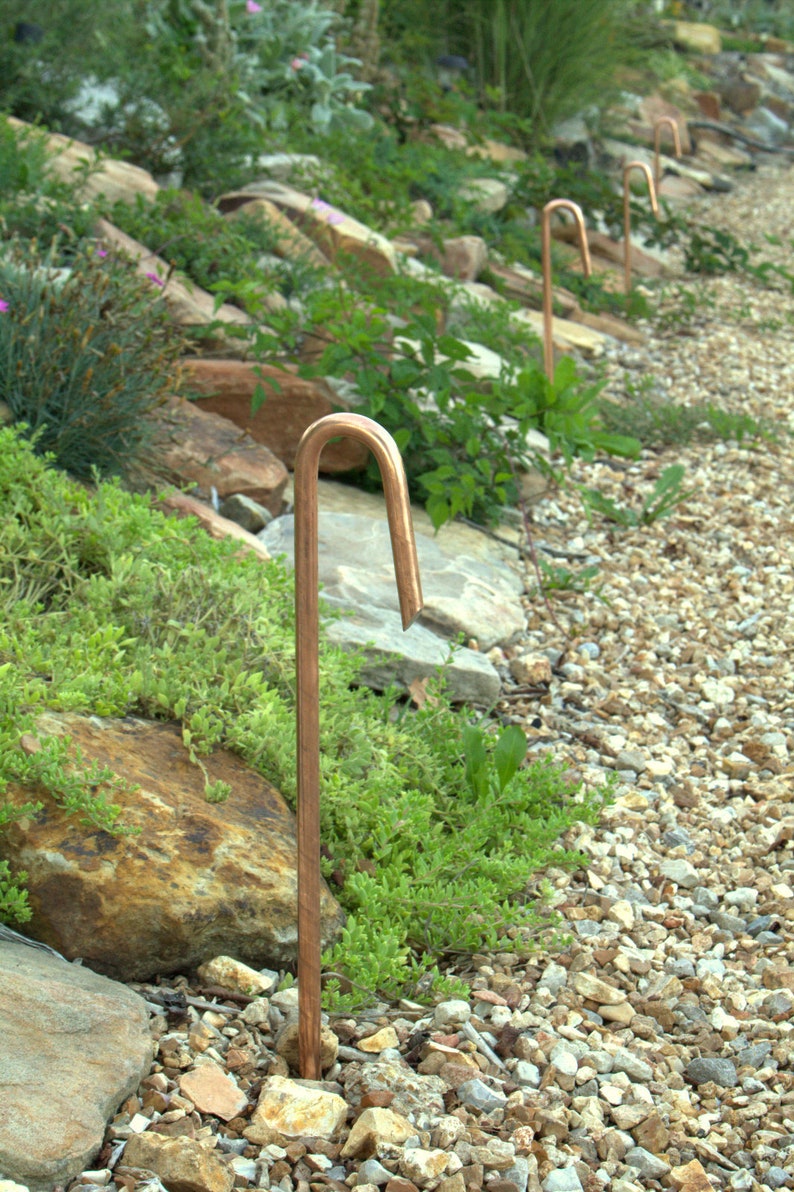 May include: A row of copper garden path lights are installed in a gravel pathway. The lights are curved at the top and are set into the ground.