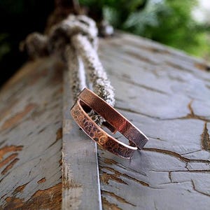 May include: Two copper bracelets with a textured, hammered design. The bracelets are a warm brown color and rest on a weathered, gray wooden surface. The background includes a blurred view of greenery and a rope.