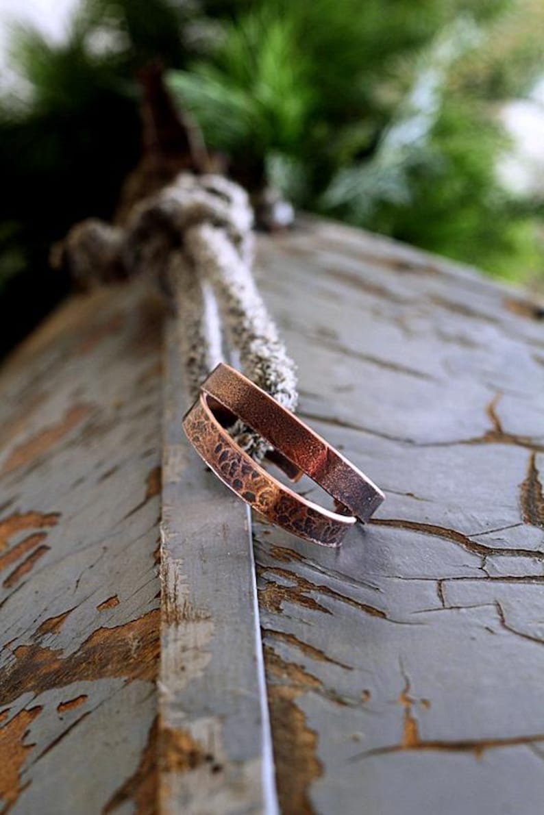 May include: Two copper bracelets with a textured, hammered design. The bracelets are a warm brown color and rest on a weathered, gray wooden surface. The background includes a blurred view of greenery and a rope.
