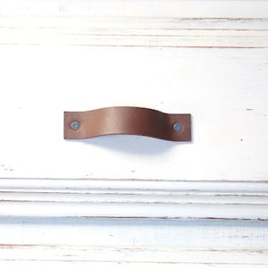 May include: A brown leather cabinet pull with a curved design, attached with two silver rivets. The pull is centered on a white, distressed wooden surface, showcasing a rustic aesthetic. This hardware is suitable for drawers and cabinets.