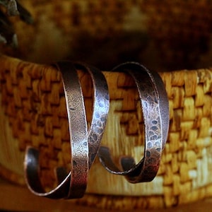 May include: Two copper-colored hoop earrings with a hammered texture are displayed against a woven basket background. The earrings have a curved, open design, showcasing a rustic, handcrafted aesthetic. The basket's natural tones complement the metal's warm hues.