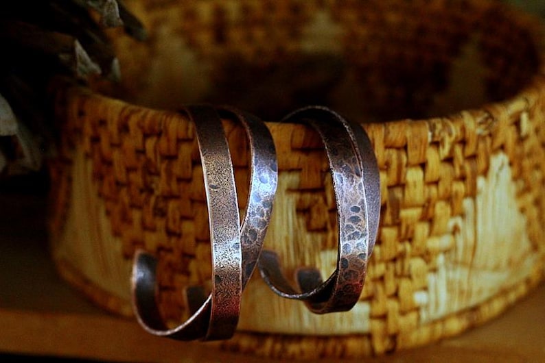 May include: Two copper-colored hoop earrings with a hammered texture are displayed against a woven basket background. The earrings have a curved, open design, showcasing a rustic, handcrafted aesthetic. The basket's natural tones complement the metal's warm hues.