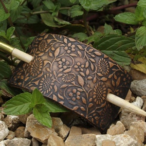 May include: A brown leather hair clip with a floral pattern and a wooden hair stick. The hair clip is on a bed of small rocks.
