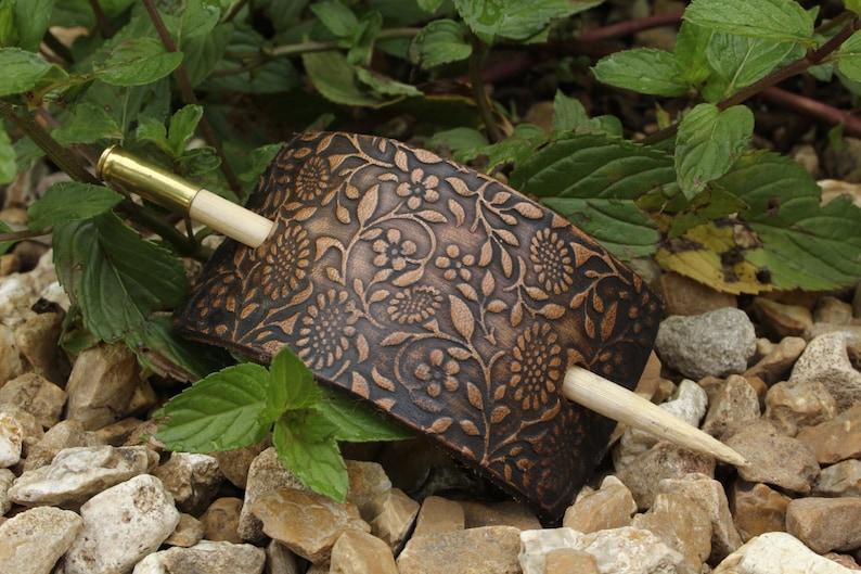 May include: A brown leather hair clip with a floral pattern and a wooden hair stick. The hair clip is on a bed of small rocks.