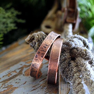 May include: Two copper cuff bracelets with textured and smooth surfaces rest on a weathered wooden surface. The bracelets have a warm, reddish-brown hue, and the textured bracelet has a hammered appearance. The background includes rope and greenery.