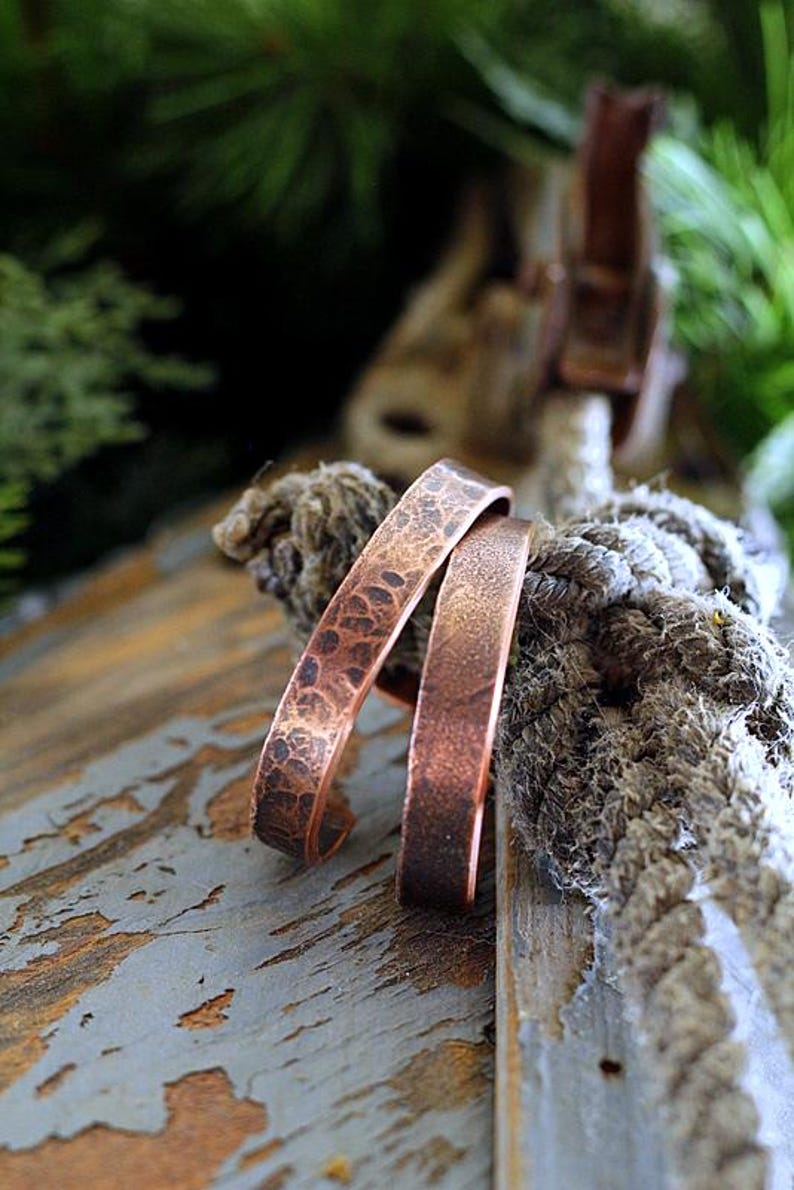 May include: Two copper cuff bracelets with textured and smooth surfaces rest on a weathered wooden surface. The bracelets have a warm, reddish-brown hue, and the textured bracelet has a hammered appearance. The background includes rope and greenery.