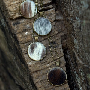 May include: Four necklaces with round pendants made of cowhide with brown and white patterns. The necklaces are hanging on a piece of weathered wood.