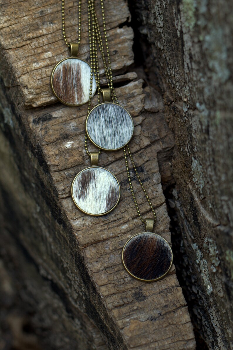 May include: Four necklaces with round pendants made of cowhide with brown and white patterns. The necklaces are hanging on a piece of weathered wood.