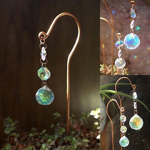 May include: A copper-colored metal plant stake with a curved top, holding a chain of iridescent crystal beads. The crystals reflect green and blue light, creating a shimmering effect. The stake is placed in a bed of small green plants.