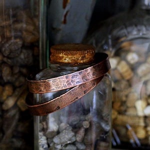 May include: A clear glass jar with a copper-colored metal band wrapped around it. The jar is filled with small, gray stones. The band has a textured surface. Other jars filled with stones are in the background.