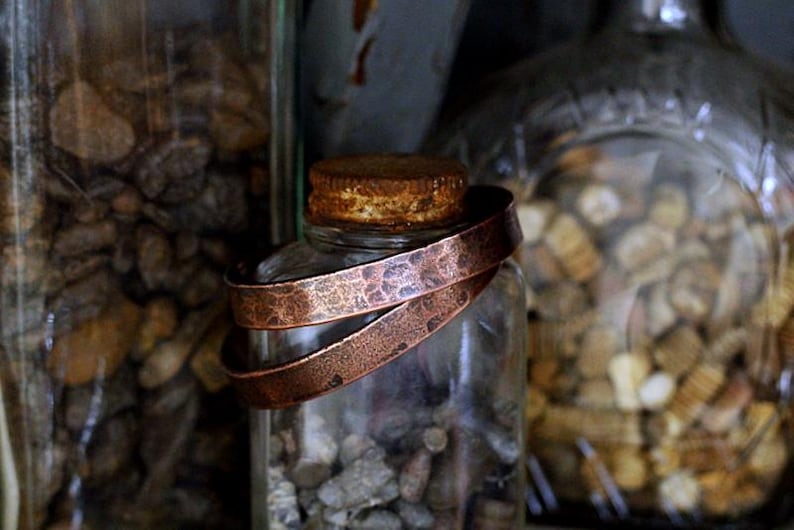 May include: A clear glass jar with a copper-colored metal band wrapped around it. The jar is filled with small, gray stones. The band has a textured surface. Other jars filled with stones are in the background.