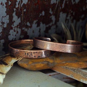 May include: Two copper cuff bracelets with textured surfaces rest on a stone. The bracelets have a hammered appearance, showcasing a rustic, handcrafted aesthetic. The warm, reddish-brown color of the copper contrasts with the neutral tones of the background.