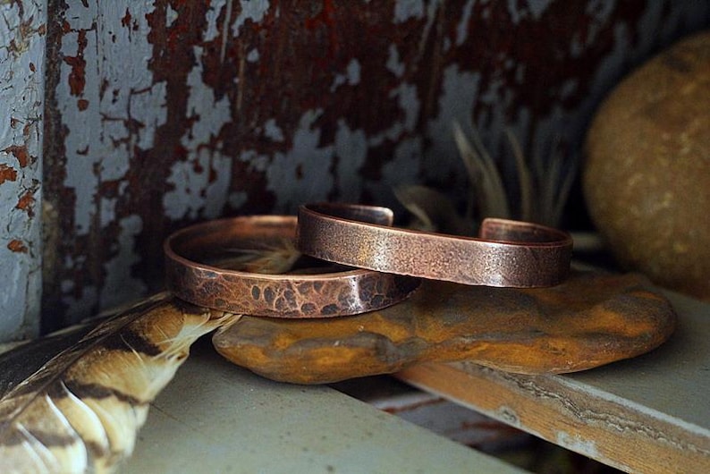 May include: Two copper cuff bracelets with textured surfaces rest on a stone. The bracelets have a hammered appearance, showcasing a rustic, handcrafted aesthetic. The warm, reddish-brown color of the copper contrasts with the neutral tones of the background.