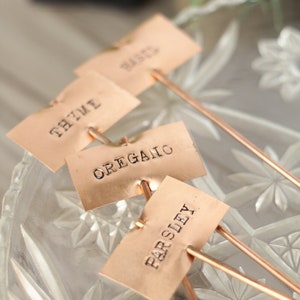May include: Four copper plant markers with the words "Thyme", "Oregano", "Parsley", and "Basil" engraved on them. The markers are on a clear glass surface.