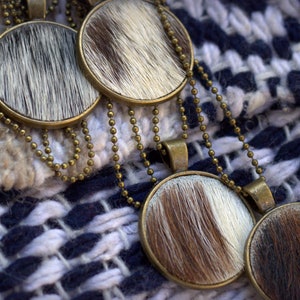 May include: Four round cowhide pendants on gold chains. The pendants are brown and white with a fuzzy texture. The chains are linked together and are lying on a white and blue woven fabric.