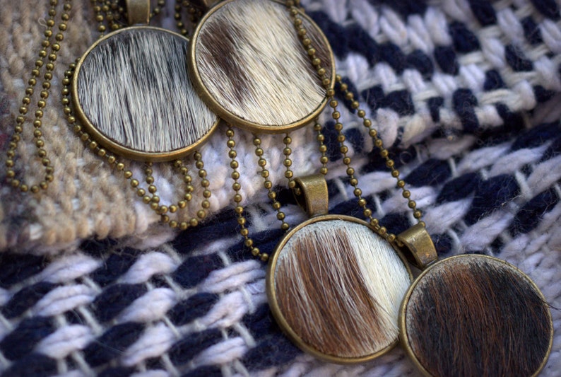 May include: Four round cowhide pendants on gold chains. The pendants are brown and white with a fuzzy texture. The chains are linked together and are lying on a white and blue woven fabric.