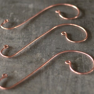 May include: Three copper-colored ornament hooks with a curved design. Each hook has a looped end for hanging and a long, straight section leading to a larger, curved section. The hooks are displayed against a gray background.