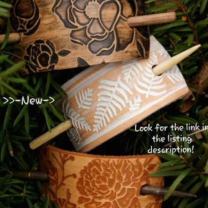 May include: Three leather hair sticks with different designs. The top stick has a floral pattern in black and brown. The middle stick has a fern pattern in white on a light brown background. The bottom stick has a floral pattern in brown on a light brown background. Each stick has a wooden dowel.