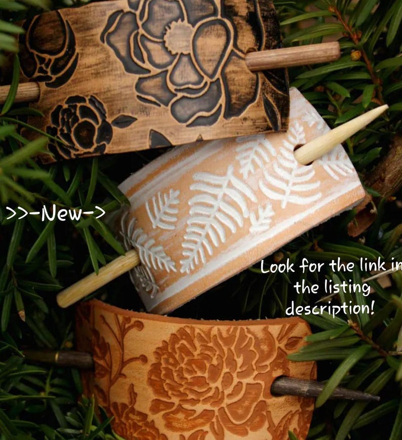 May include: Three leather hair sticks with different designs. The top stick has a floral pattern in black and brown. The middle stick has a fern pattern in white on a light brown background. The bottom stick has a floral pattern in brown on a light brown background. Each stick has a wooden dowel.