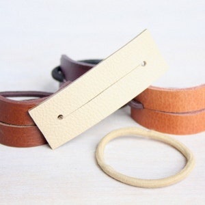 Op de afbeelding: Drie leren haar elastiekjes, een bruin, een beige en een donkerbruin. Het beige haar elastiekje heeft een cr&egrave;mekleurig leren bandje met twee gaatjes.