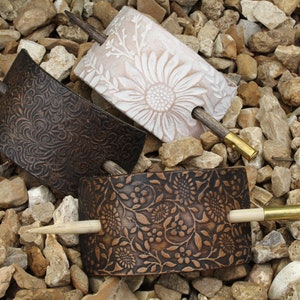 May include: Three hair clips made of leather with different floral patterns. Each clip has a wooden stick with a brass tip.