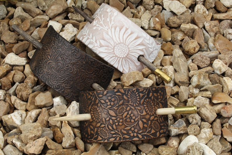 May include: Three hair clips made of leather with different floral patterns. Each clip has a wooden stick with a brass tip.