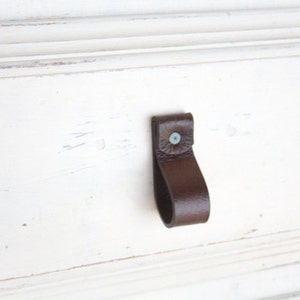 May include: A dark brown leather drawer pull with a loop design, attached to a white painted wooden surface. The pull is secured with a small, light-colored rivet.