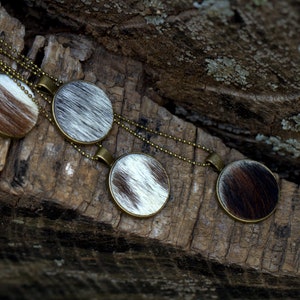May include: Four round pendants with a cowhide pattern hang on a gold chain. The pendants are on a rustic wooden surface.