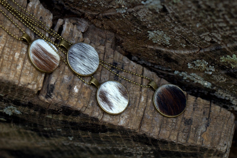 May include: Four round pendants with a cowhide pattern hang on a gold chain. The pendants are on a rustic wooden surface.