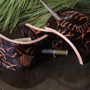 Puede incluir: Dos accesorios para el cabello de cuero marr&oacute;n oscuro con intrincados dise&ntilde;os florales y palos de madera. El cuero est&aacute; grabado con un dise&ntilde;o detallado y tiene un color marr&oacute;n m&aacute;s claro que resalta el patr&oacute;n. Los accesorios se exhiben sobre una superficie de madera oscura.
