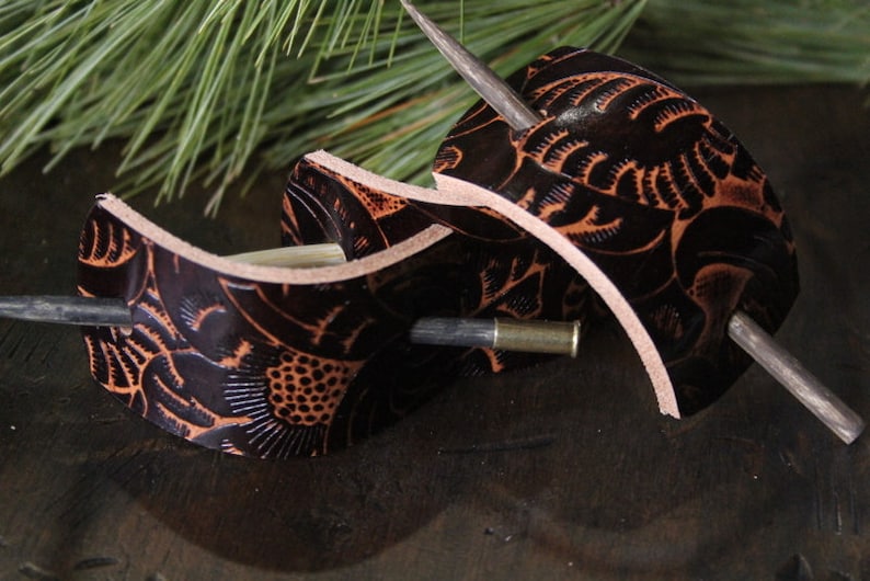 Puede incluir: Dos accesorios para el cabello de cuero marr&oacute;n oscuro con intrincados dise&ntilde;os florales y palos de madera. El cuero est&aacute; grabado con un dise&ntilde;o detallado y tiene un color marr&oacute;n m&aacute;s claro que resalta el patr&oacute;n. Los accesorios se exhiben sobre una superficie de madera oscura.
