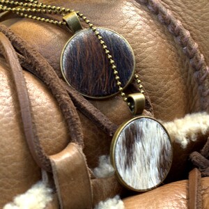 May include: Two round pendants with a brown and white cowhide pattern hang from a gold chain on a brown leather background.