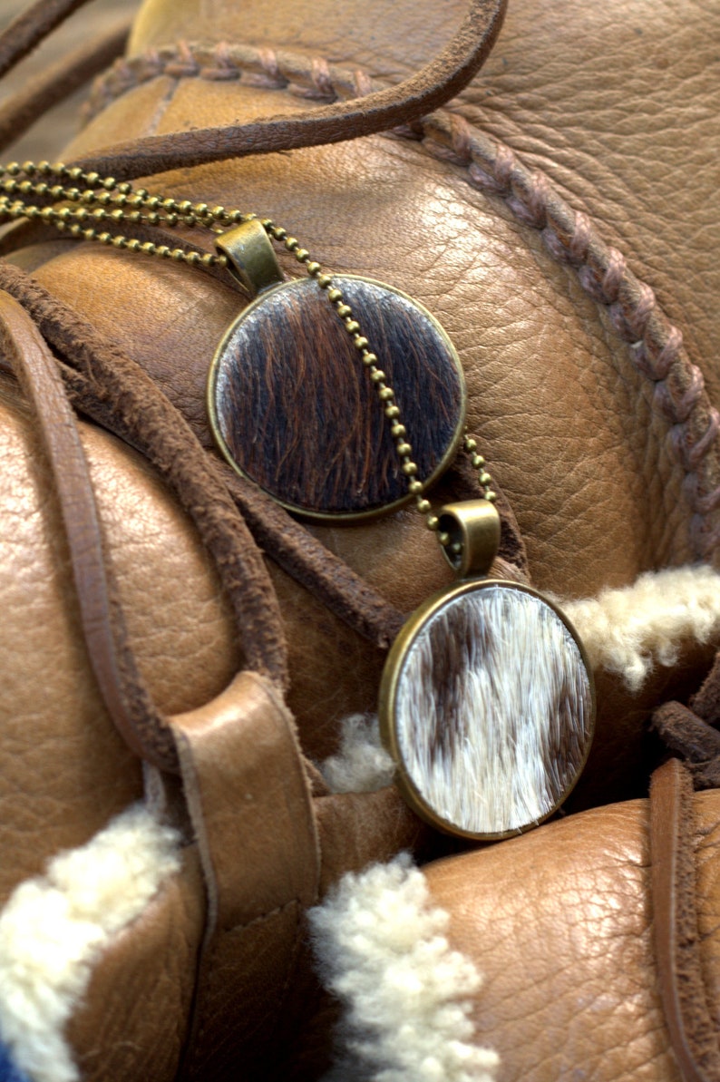 May include: Two round pendants with a brown and white cowhide pattern hang from a gold chain on a brown leather background.