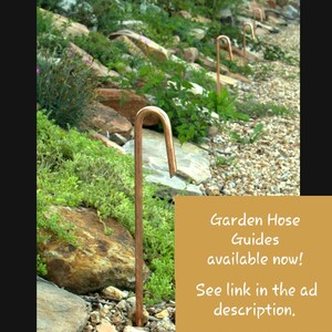 May include: A row of copper garden hose guides are installed in a rock garden. The guides are shaped like a shepherd's crook. The text in the image reads "Garden Hose Guides available now! See link in the ad description."