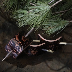 Puede incluir: Tres horquillas de cuero marr&oacute;n oscuro con intrincados dise&ntilde;os tallados y clavijas de madera. Los accesorios para el cabello est&aacute;n dispuestos sobre una superficie oscura y texturizada con agujas de pino en el fondo.