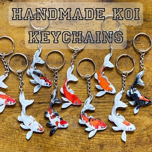 May include: A collection of nine koi fish keychains with metal rings and chains. Each keychain features a detailed illustration of a koi fish in various color patterns including orange, white, black, and red. The text HANDMADE KOI KEYCHAINS is visible.