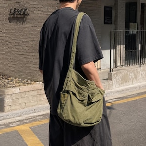 May include: An olive green canvas crossbody bag featuring a wide shoulder strap, a large main compartment, and two front pockets with flap closures. The bag is worn over the shoulder, resting against the side of a dark gray t-shirt and black pants.