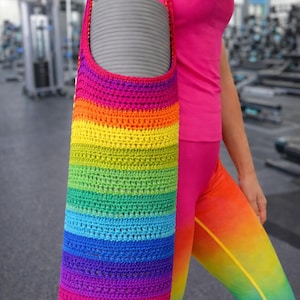 May include: A rainbow-colored crochet yoga mat bag. The bag is cylindrical, with a strap for carrying. The colors transition from red to purple, green, and yellow. A gray yoga mat is inside the bag. The background is a gym.