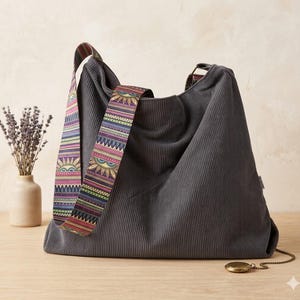 May include: A gray corduroy shoulder bag with a colorful patterned strap featuring sun and geometric designs. The bag has a large capacity and a simple design. A small, round, gold-colored locket with a chain rests on the table.