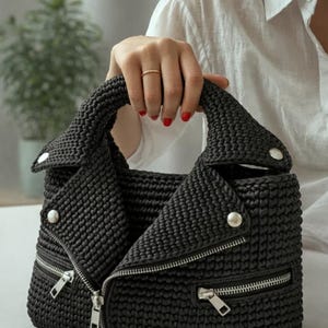 Puede incluir: Un bolso negro de ganchillo con forma de chaqueta de motorista. El bolso tiene cierre de cremallera, herrajes plateados y un aspecto tejido texturizado. El bolso lo sostiene una persona con esmalte de uñas rojo.