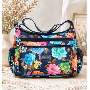 May include: A navy blue crossbody bag featuring a vibrant floral pattern with pink, orange, yellow, light blue, and green flowers. The bag has multiple zippered compartments, side pockets, an adjustable shoulder strap, and a metal logo plate reading RUIMOTI XIUXIANXILIE.