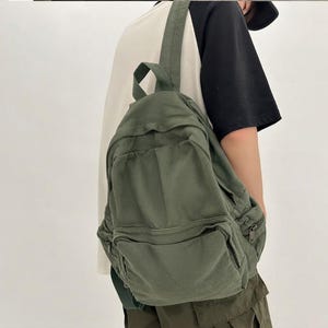 May include: An olive green canvas backpack featuring a large main compartment, a smaller front zippered pocket, and matching olive green shoulder straps. The backpack has a simple, functional design with visible zipper closures and fabric construction.