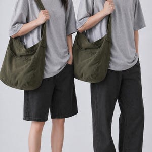 Pode incluir: Duas pessoas vestindo camisetas cinza e calças pretas, cada uma carregando uma grande bolsa de ombro verde oliva. As bolsas têm uma alça larga e um bolso grande. O fundo é branco liso.