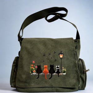 May include: An olive green canvas messenger bag featuring a graphic print of four cats sitting on a ledge next to a street lamp. The cats are black, orange, black, and black-and-white. Small birds fly above the cats, and orange flowers grow nearby.