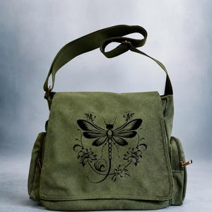 May include: An olive green canvas messenger bag featuring a black graphic print of a dragonfly surrounded by floral elements on the front flap. The bag includes an adjustable shoulder strap and side pockets with metallic zippers on both sides.