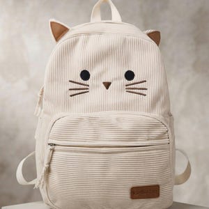 May include: A cream-coloured corduroy backpack featuring a cat face design with embroidered black eyes, a brown triangular nose, and brown whiskers. The bag includes two small brown ears on top, a front zippered pocket, and a brown brand label.