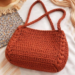May include: A rust-coloured crochet bag with braided handles and edges. The bag has a textured stitch pattern and a rectangular shape. The handles are thick and sturdy, designed for comfortable carrying. The bag is a stylish accessory.